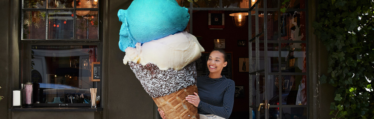 Eine Frau steht lächelnd vor einem Gebäude und hält eine überdimensionale Eiswaffel mit drei Kugeln in Blau, Weiß und Braun.