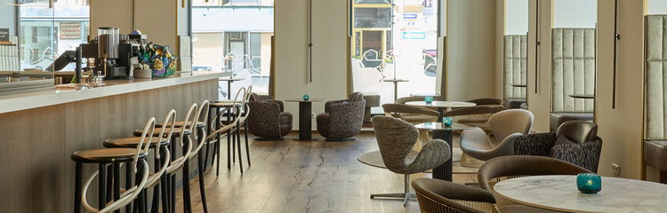 Elegantes Hotelcafé mit Bar, hohen Stühlen und gemütlichen Sitzgruppen, hellem Tageslicht und großer Fensterfront, die eine einladende Atmosphäre für entspanntes Frühstück oder Kaffee bietet.